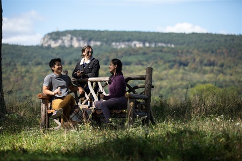 Three people sitting on a rustic bench, drinking beer and wine, with a view of Skytop Tower on the Shawangunk Ridge in the background.